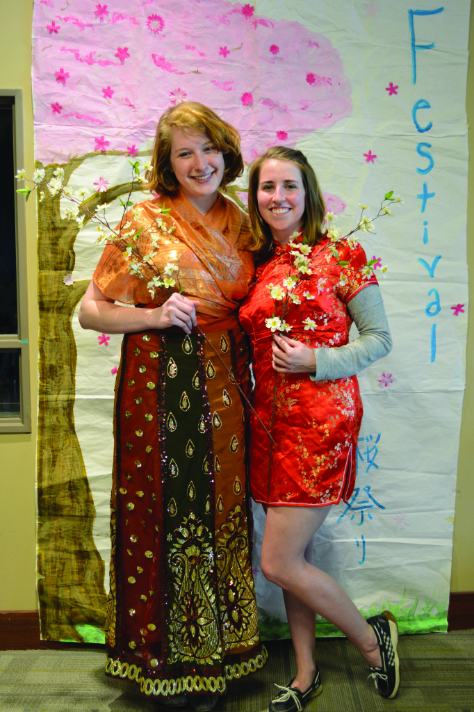 Maddy Bellman ‘18 and a friend pose in front of student artwork while holding cherry blossom flowers.